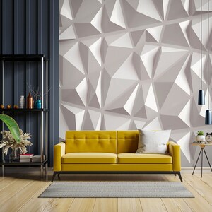 May include: A yellow sofa with a white pillow sits in front of a wall with a geometric pattern. The wall is a light gray color with white geometric shapes. The sofa is in a living room with a wooden floor and a black shelf unit in the background.