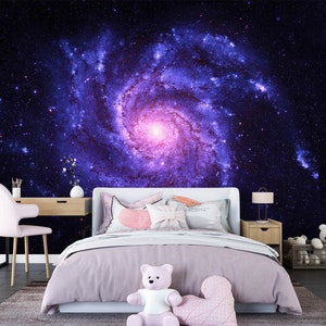 May include: A bedroom with a galaxy-themed mural. The mural features a spiral galaxy in shades of purple and pink against a dark blue background. The room includes a bed with pink bedding, a desk, a chair, and decorative items.