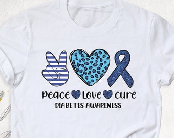 Puede incluir: Camiseta blanca con un signo de paz azul, un corazón azul con estampado de leopardo y una cinta azul. El texto "peace love cure" está debajo de los símbolos, y el texto "diabetes awareness" está debajo de ese.