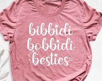 May include: A pink t-shirt with the text "bibbidi bobbidi besties" in white lettering with stars and a heart.