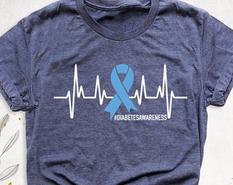 Puede incluir: Una camiseta azul oscuro con un gráfico de latido del corazón blanco y una cinta azul. El texto "#DIABETESAWARENESS" está impreso debajo del gráfico.