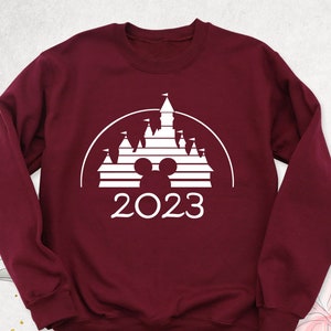 May include: Burgundy sweatshirt with a white graphic of the Disney castle, Mickey Mouse ears, and the year 2023.