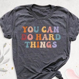 May include: A dark gray t-shirt with the text "YOU CAN DO HARD THINGS" in rainbow colors.