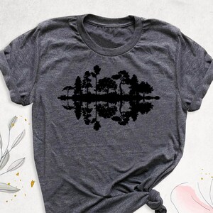May include: A dark gray t-shirt with a black silhouette of trees reflected in water. The design is centered on the shirt.