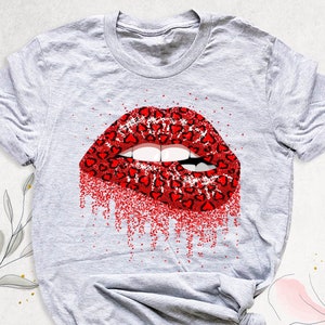 May include: A light gray t-shirt featuring a graphic of red lips with a leopard print pattern of hearts. The lips are partially biting the lower lip, with red glitter dripping down. The shirt is a casual style.