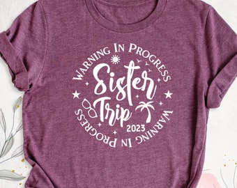 May include: A purple t-shirt with a white circle graphic that says "Warning in Progress Sister Trip 2023 Warning in Progress".