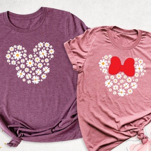 May include: Two t-shirts with a daisy flower design in the shape of Mickey Mouse and Minnie Mouse. The Mickey Mouse shirt is purple and the Minnie Mouse shirt is pink.