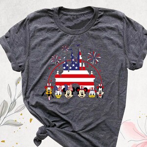 May include: A gray t-shirt with a graphic of Mickey Mouse, Minnie Mouse, Donald Duck, Goofy, and Pluto in front of a castle with an American flag. The characters are wearing red, white, and blue hats. There are fireworks in the background.
