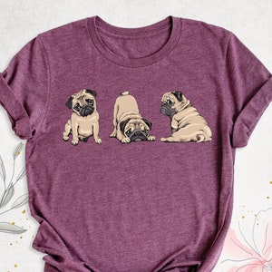May include: A purple t-shirt with a graphic of three pugs. The pugs are in different poses, one is sitting, one is looking at the camera, and one is laying down.