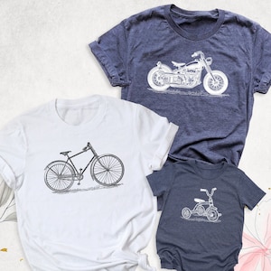 May include: Three t-shirts featuring different vehicle designs. The largest t-shirt is navy blue with a white motorcycle design. The medium t-shirt is white with a black bicycle design. The smallest t-shirt is navy blue with a white tricycle design.