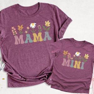 May include: Two matching maroon t-shirts. The larger shirt has the word "MAMA" in a retro font with a floral design above it. The smaller shirt has the word "MINI" with a similar floral design.