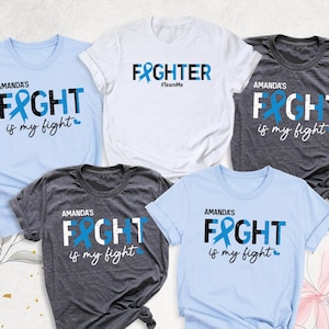May include: Five t-shirts with different designs. Two are light blue, two are dark gray, and one is white. The designs feature the words "Amanda's Fight is my fight" with a blue ribbon, and "Fighter #TeamMe".