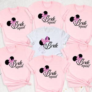 May include: Six pink and white t-shirts with a black and white image of Mickey Mouse ears with a pink bow and the text "Bride Squad" on each shirt.