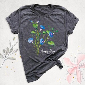 May include: Dark gray t-shirt featuring a watercolor illustration of blue morning glory flowers with green leaves and vines. The text "Morning Glory" is written in white script below the flowers.