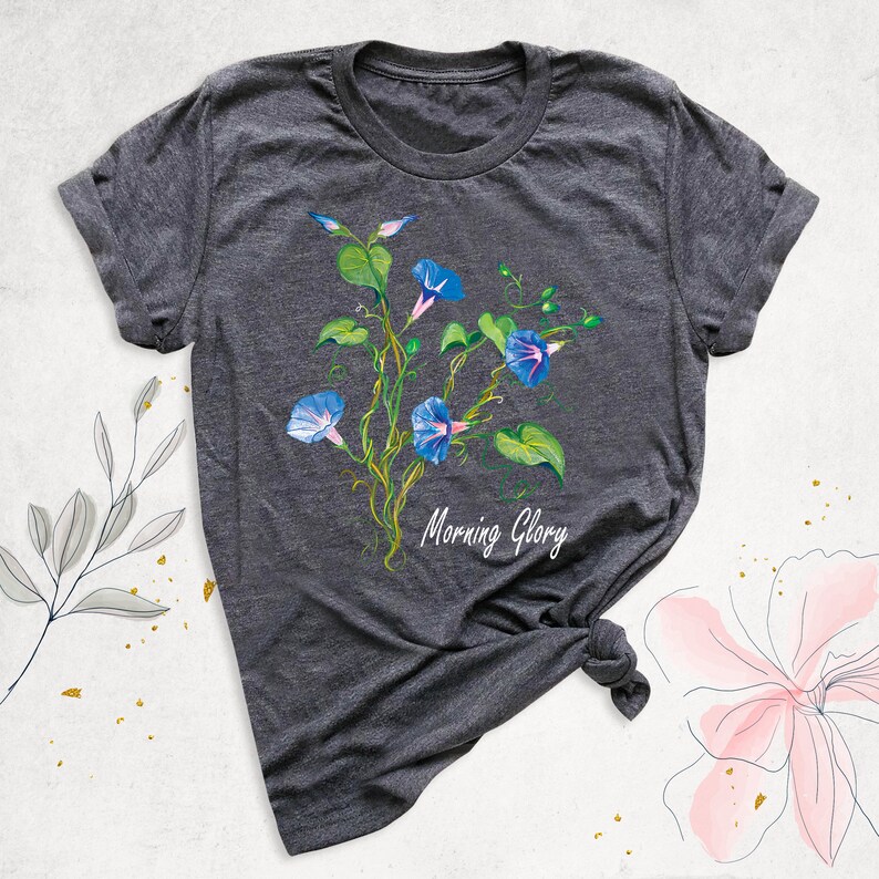May include: Dark gray t-shirt featuring a watercolor illustration of blue morning glory flowers with green leaves and vines. The text "Morning Glory" is written in white script below the flowers.