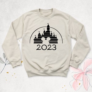 May include: A beige sweatshirt with a black silhouette of the Cinderella Castle from Disney World. The castle has a Mickey Mouse head in the center and the year 2023 below it.