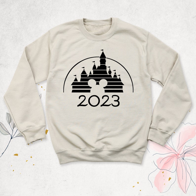 May include: A beige sweatshirt with a black silhouette of the Cinderella Castle from Disney World. The castle has a Mickey Mouse head in the center and the year 2023 below it.