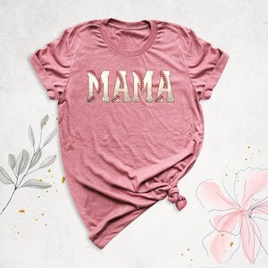 May include: A pink t-shirt with the word "MAMA" written in white letters with a baseball stitch design.