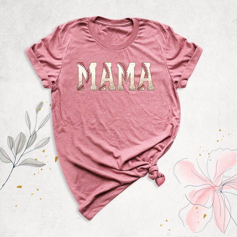 May include: A pink t-shirt with the word "MAMA" written in white letters with a baseball stitch design.