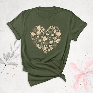 May include: An olive green short-sleeved t-shirt featuring a central graphic design composed of various beige seashells, starfish, and coral arranged in the shape of a heart. The shirt has a crew neckline and cuffed sleeves.