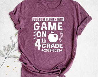 May include: A heathered maroon short-sleeve t-shirt featuring white text that reads CUSTOM ELEMENTARY GAME ON 4th GRADE 2022-2023, accompanied by a graphic of an apple with a heart cutout and a pencil icon centred on the front.