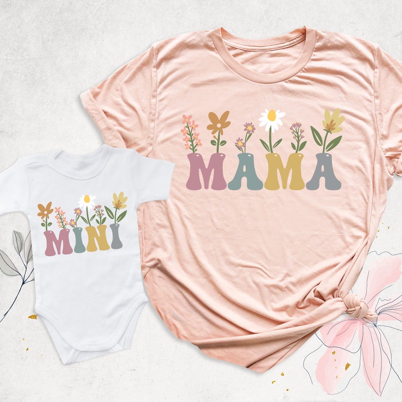 May include: A light pink t-shirt and a white baby onesie. Both feature the word "MAMA" and "MINI" in colorful letters, with floral arrangements in vases above the text. The t-shirt is knotted at the bottom.