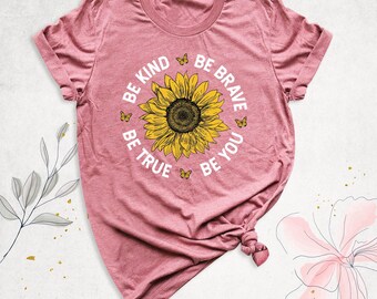 May include: A heathered mauve short-sleeve t-shirt featuring a large yellow sunflower graphic in the center. Surrounding the sunflower are small yellow butterflies and the white text BE KIND BE BRAVE BE TRUE BE YOU in a circular layout.