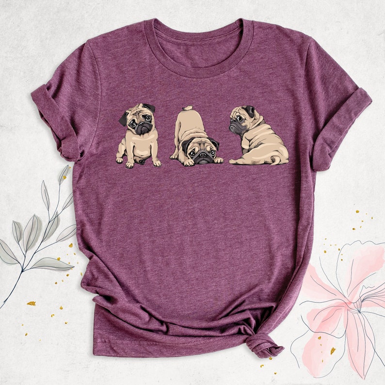 May include: A purple t-shirt with a graphic of three pugs. The pugs are in different poses, one is sitting, one is looking at the camera, and one is laying down.