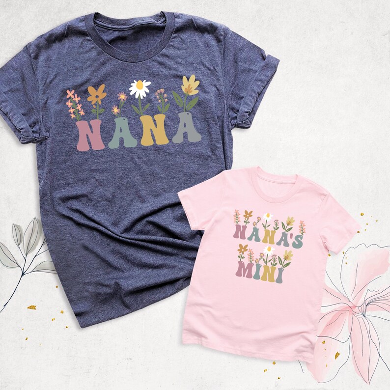 May include: Two t-shirts, one navy blue and one pink, with floral designs. The navy shirt says "NANA" in colorful letters with flowers above. The pink shirt says "NANA'S MINI" with flowers. Both shirts have short sleeves.