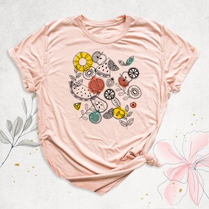 May include: A pink t-shirt with a graphic design of various fruits and leaves in black, yellow, red, and green. The design includes watermelon, pineapple, kiwi, lemons, and apples.