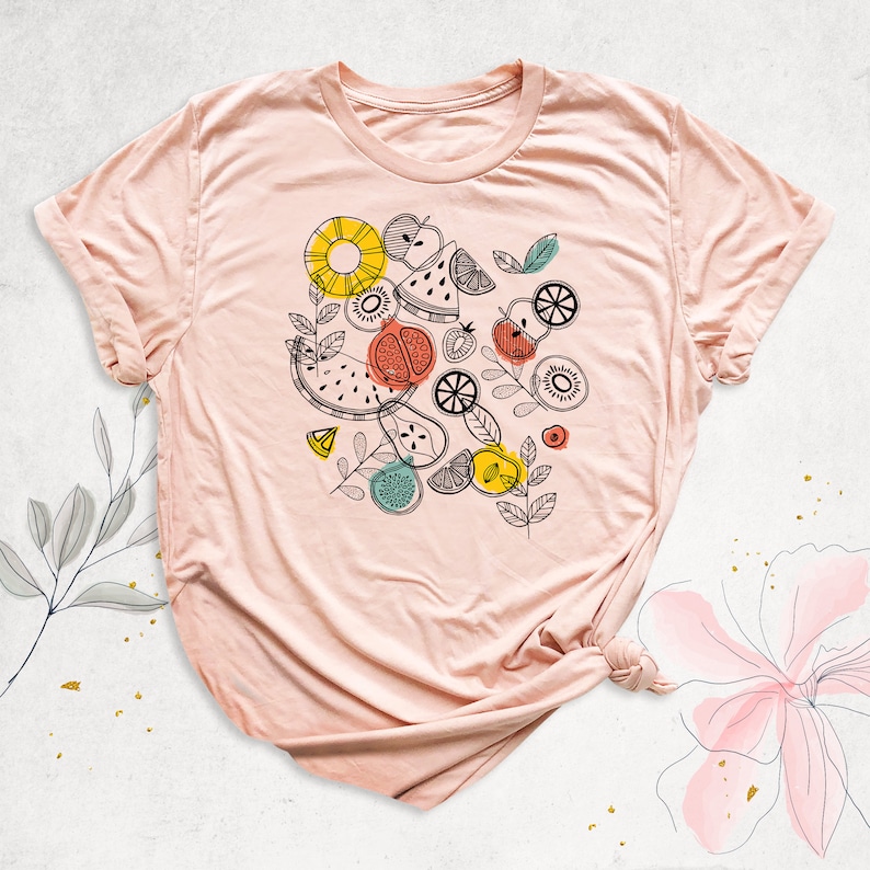 May include: A pink t-shirt with a graphic design of various fruits and leaves in black, yellow, red, and green. The design includes watermelon, pineapple, kiwi, lemons, and apples.