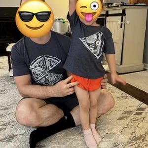以下が含まれることがあります： チャコールグレーの半袖Tシャツ2枚。白いピザのスライスのグラフィックプリントが施されています。大きい方のTシャツは胸元にピザのグラフィックがあり、小さい方のTシャツは胸の上部に同じピザのスライスのグラフィックがデザインされています。
