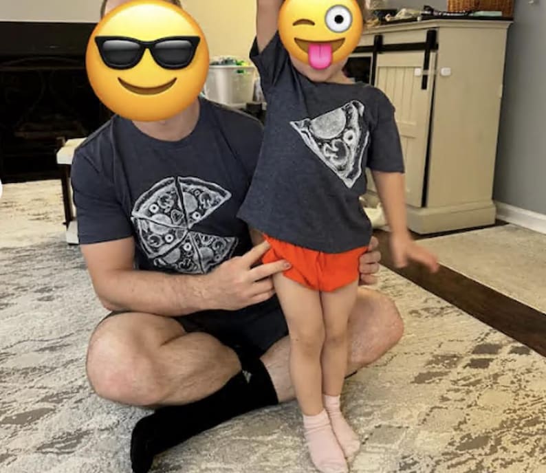 以下が含まれることがあります： チャコールグレーの半袖Tシャツ2枚。白いピザのスライスのグラフィックプリントが施されています。大きい方のTシャツは胸元にピザのグラフィックがあり、小さい方のTシャツは胸の上部に同じピザのスライスのグラフィックがデザインされています。