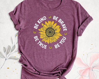 May include: A heathered maroon short-sleeve t-shirt featuring a central yellow sunflower graphic surrounded by small yellow butterflies and the white text BE KIND BE BRAVE BE TRUE BE YOU arranged in a circular pattern around the flower.