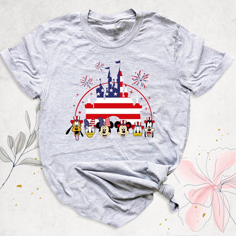 May include: A gray t-shirt with a graphic of Mickey Mouse, Minnie Mouse, Donald Duck, Goofy, and Pluto in front of a castle with an American flag. The castle is surrounded by fireworks.