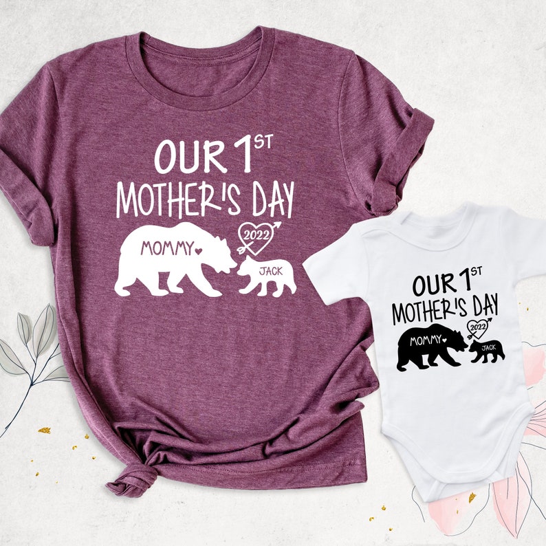 Puede incluir: Un conjunto de dos camisetas, una para un padre y otra para un ni&ntilde;o, celebrando su primer D&iacute;a de la Madre. La camiseta para adultos es una camiseta color marr&oacute;n con el texto "OUR 1ST MOTHER'S DAY" y un gr&aacute;fico de dos osos, uno m&aacute;s grande que el otro. El oso m&aacute;s peque&ntilde;o est&aacute; etiquetado como "JACK" y el oso m&aacute;s grande est&aacute; etiquetado como "MOMMY". Los osos se miran y el oso m&aacute;s grande tiene un coraz&oacute;n con el a&ntilde;o "2022" dentro. La camiseta para ni&ntilde;os es un mono blanco con el mismo texto y gr&aacute;fico que la camiseta para adultos.