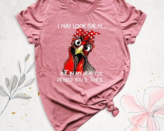 May include: A heathered mauve short-sleeve t-shirt featuring a graphic of a chicken wearing red-rimmed glasses and a red polka-dot headscarf. The text reads: I MAY LOOK CALM... BUT IN MY HEAD I'VE PECKED YOU 3 TIMES...