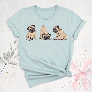 May include: A light blue t-shirt with a graphic of three pugs in different poses. The pugs are brown and white with black noses and eyes.