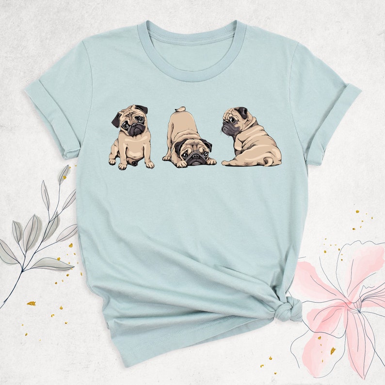 May include: A light blue t-shirt with a graphic of three pugs in different poses. The pugs are brown and white with black noses and eyes.