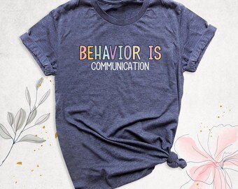 May include: A heather navy blue short-sleeve t-shirt featuring the text "BEHAVIOR IS COMMUNICATION" printed in a multi-colored, pastel-toned font across the chest. The shirt has a crew neckline and a standard fit with folded sleeves.