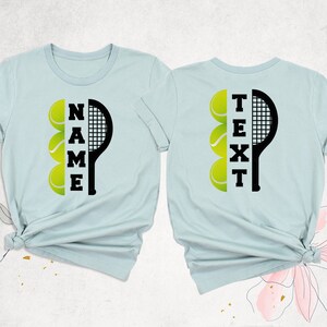 May include: Two light blue t-shirts with a black tennis racket and yellow tennis balls. The text "NAME" is on the left shirt and "TEXT" is on the right shirt.