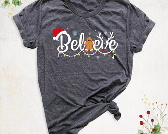 May include: A dark heather gray short-sleeve t-shirt featuring the white text "Believe" with a red Santa hat on the B, a gingerbread man replacing the letter i, and reindeer antlers on the e, accented by colorful string lights and snowflakes.