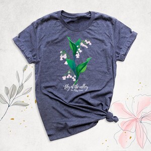May include: A dark heather grey t-shirt with a white graphic of a lily of the valley flower. The text "Lily of the valley, the May flower" is printed below the flower.