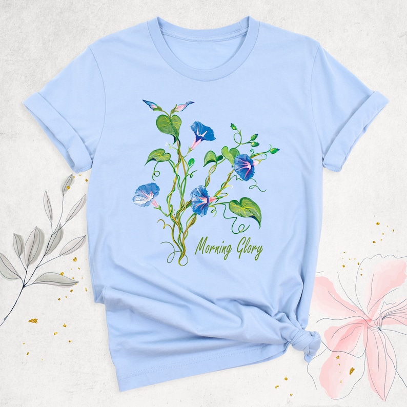 May include: Light blue t-shirt featuring a watercolor illustration of morning glory flowers. The design includes blue flowers, green leaves, and the text "Morning Glory" in green script. The shirt has rolled-up sleeves.