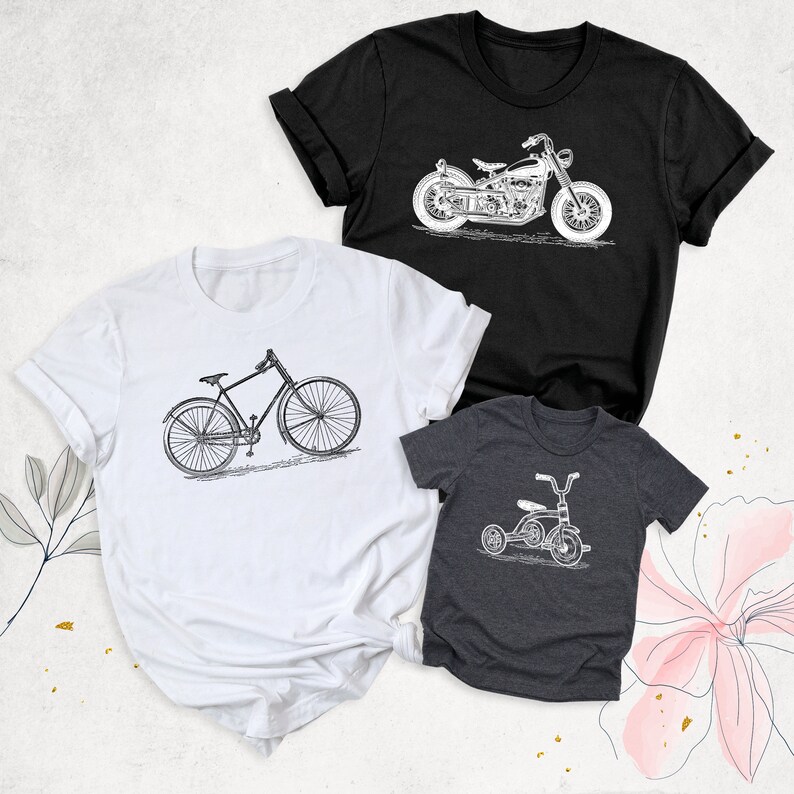 May include: Three t-shirts featuring different vintage vehicle designs. A white t-shirt with a bicycle, a black t-shirt with a motorcycle, and a grey t-shirt with a tricycle.