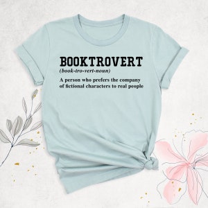 May include: A light blue t-shirt with the text "BOOKTROVERT (book-tro-vert-noun) A person who prefers the company of fictional characters to real people" printed in black.