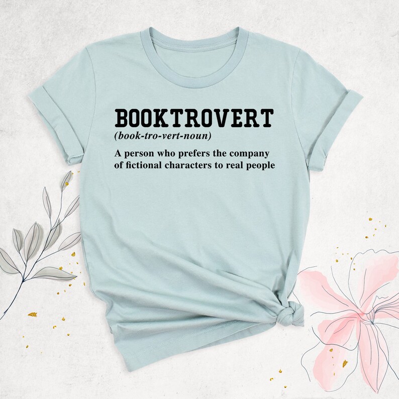 May include: A light blue t-shirt with the text "BOOKTROVERT (book-tro-vert-noun) A person who prefers the company of fictional characters to real people" printed in black.