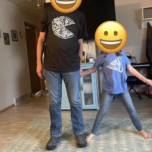 以下が含まれることがあります： 一切れ欠けたピザの白いグラフィックが描かれた黒い半袖Tシャツと青いデニムジーンズ、グレーのスニーカー、そしてピザ一切れの白いグラフィックが描かれた水色の半袖Tシャツ。