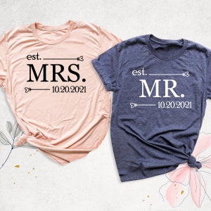 K&ouml;nnte beinhalten: Zwei T-Shirts, eines rosa und eines blau, mit dem Text "est. MRS. 10.20.2021" und "est. MR. 10.20.2021" bedruckt. Die Shirts sind gefaltet und auf einem floralen Hintergrund platziert.