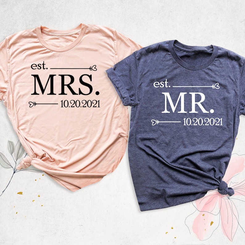K&ouml;nnte beinhalten: Zwei T-Shirts, eines rosa und eines blau, mit dem Text "est. MRS. 10.20.2021" und "est. MR. 10.20.2021" bedruckt. Die Shirts sind gefaltet und auf einem floralen Hintergrund platziert.
